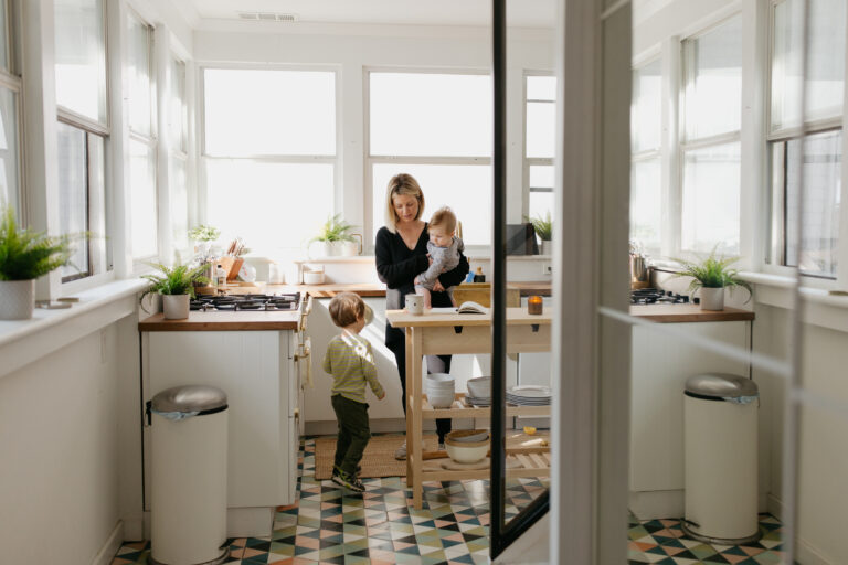 A mother holds her young child in a kitchen and a toddler walks towards her