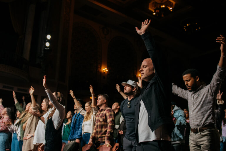 A crowd of worshippers stands with hands raised in praise